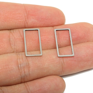 Puede incluir: Dos componentes rectangulares plateados para pendientes. Cada componente es un rectángulo hueco con un marco delgado. Los componentes se muestran en una mano, con un fondo blanco.