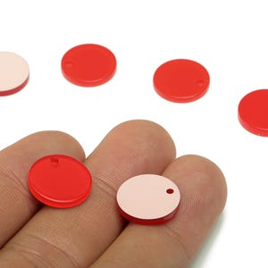Pode incluir: Pingentes redondos de acrílico vermelho e rosa com um furo para fazer joias. Os pingentes estão em vários tamanhos e são mostrados em um fundo branco. Alguns pingentes são vermelhos e outros rosa. Os pingentes estão sendo segurados em uma mão.