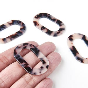 Peut inclure: Quatre maillons ovales en résine avec un motif écaille de tortue crème, marron et noir. Un maillon est tenu dans une main, les autres sont dispersés sur une surface blanche. Chaque maillon a un petit trou en haut.