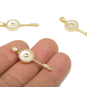May include: Gold-colored key pendant with a heart-shaped design. The key features a round head with a heart-shaped inlay, surrounded by small, clear stones. The key's stem is detailed with small lines and a decorative end.