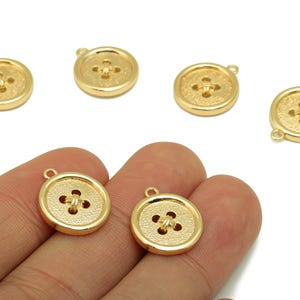 Könnte beinhalten: Goldfarbene Knopfanhänger mit klassischem Vier-Loch-Design. Die Knöpfe haben eine strukturierte Oberfläche und eine kleine Schlaufe zum Befestigen an Schmuck oder anderen Bastelarbeiten. Mehrere Knöpfe sind auf weißem Hintergrund verstreut.