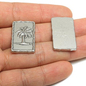 Puede incluir: Dos dijes rectangulares de metal plateado. Un dije presenta un diseño detallado de palmera dentro de un borde texturizado. El otro dije es un rectángulo liso. Estos dijes son probablemente para la fabricación de joyas o manualidades.