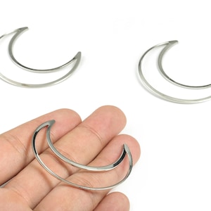 Puede incluir: Encantos de luna creciente de metal plateado. Los amuletos tienen un diseño de doble capa, con un acabado liso y pulido. La imagen muestra tres amuletos, uno de ellos sostenido en una mano. Estos amuletos son adecuados para la fabricación de joyas y la artesanía.