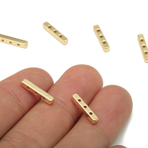 May include: A collection of small, rectangular gold-coloured metal beads, each with three holes, are shown against a white backdrop. Two beads are held in a hand. These beads are likely for jewellery making or craft projects.