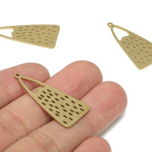 Puede incluir: Tres colgantes geométricos de color latón. Cada colgante tiene forma triangular con la parte superior redondeada y una serie de pequeños recortes ovalados espaciados uniformemente. Un colgante se sostiene en una mano.