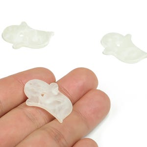 Könnte beinhalten: Drei cremefarbene, geisterförmige Bastelperlen mit Marmoreffekt. Eine Perle wird in einer Hand gehalten, während die anderen beiden auf weißem Hintergrund verstreut sind. Die Perlen haben ein Loch zum Auffädeln.