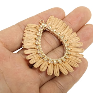 Op de afbeelding: Een goudkleurige, druppelvormige hanger met een geweven zonnestraalontwerp. De hanger heeft een centrale open ruimte en is versierd met kleine, heldere kristallen langs de binnenrand. De hanger wordt in een hand gehouden.
