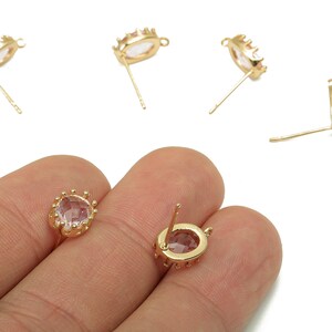 May include: Gold-coloured stud earrings with a crown-like design. Each earring features a light pink oval-shaped stone. The earrings are shown on a white background, with some held in a hand.