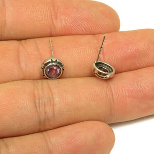 Könnte beinhalten: Ein Paar Ohrstecker mit silberfarbener Fassung und einem runden, hellvioletten Edelstein. Die Ohrringe werden auf einer Hand präsentiert, die die filigranen Details des Schmucks zeigt.