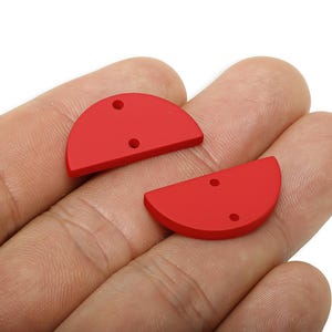 May include: Two bright red, semi-circular pendants with two small holes each. The pendants are displayed against a neutral background, suggesting they are jewelry components or craft supplies. The pendants are made of a solid material.