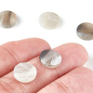 Puede incluir: Un conjunto de cuentas redondas veteadas en tonos grises y blancos. Las cuentas son lisas y pulidas, con un patrón arremolinado. Algunas cuentas se sostienen en una mano, mientras que otras están sobre una superficie blanca. Estas cuentas son adecuadas para la fabricación de joyas.