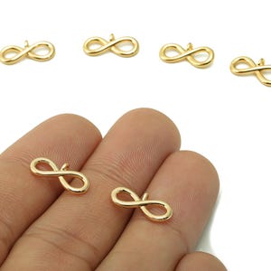 May include: Gold-colored infinity charm pendants. The charms are in the shape of the infinity symbol, with a small loop at the top for attaching to jewelry. The charms are shown on a white background and in a person's hand.