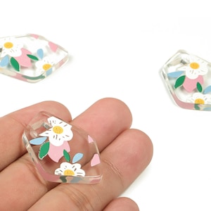 Op de afbeelding: Transparante, onregelmatig gevormde acryl bedels met een bloemenontwerp. Elke bedel heeft witte bloemen met gele centra, roze bloemblaadjes en groene en blauwe bladeren. Ideaal voor het maken van sieraden.