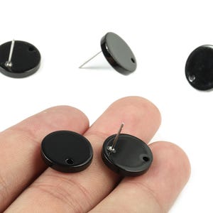 May include: Black, round stud earrings with a glossy finish. The earrings have a post and a small hole for the backing. Several earrings are shown, some with posts visible, and some held in a hand.