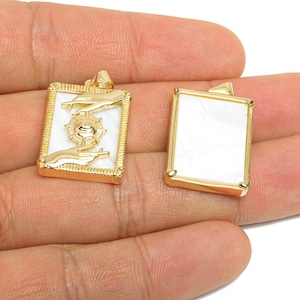 May include: Two gold-colored rectangular pendants. One pendant features a white background with a gold-colored design of hands and a wheel. The other pendant has a white background. Both pendants have a gold-colored frame.