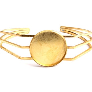 May include: A gold metal bracelet with a round, empty, gold-toned bezel. The bracelet is made of three bands of metal that are connected together.