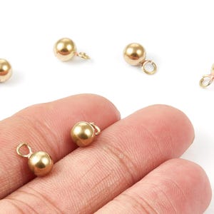 May include: Close-up of several small, gold-colored ball charms with attached loops. The charms are scattered on a white background, with some resting on a person's hand. These jewelry findings are ideal for crafting.