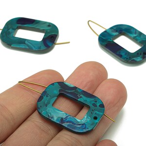 Puede incluir: Pendientes rectangulares de color turquesa y azul marino con efecto marmolado. Los pendientes tienen un recorte rectangular en el centro y están suspendidos de alambres dorados. Se muestra en una mano.