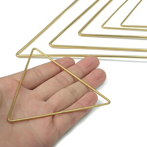 Könnte beinhalten: Eine Hand hält ein kleines, goldfarbenes Dreieck. Mehrere größere, ähnliche Dreiecke sind im Hintergrund angeordnet. Die Dreiecke scheinen aus dünnem Draht oder Metall zu bestehen, vor weißem Hintergrund.