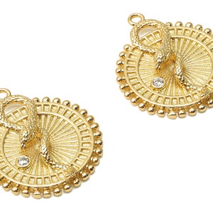 以下が含まれることがあります： 蛇のデザインと中央のクリアなラインストーンが特徴の、ゴールドトーンのペンダントチャーム2個。各チャームは小さなビーズの円に囲まれています。