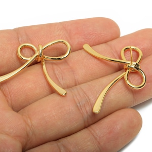 May include: Two gold-colored bow-shaped jewelry findings. Each bow has a looped center and curved, elongated ends. The findings are displayed against a neutral background, highlighting their shiny, metallic finish. These are likely used for jewelry making.