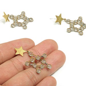 May include: Gold-toned star-shaped earrings with a geometric design. Each earring features a small star charm and a larger star-shaped structure with clear, round accents. The earrings are displayed against a white background.