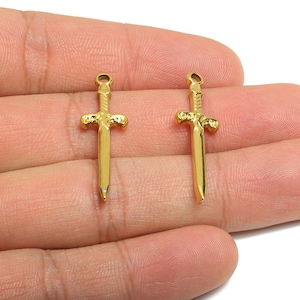 May include: Two small, gold-coloured sword pendants. The pendants are held in a person's hand. Each sword has a detailed hilt and a pointed blade. The pendants are likely for jewellery making or crafting.