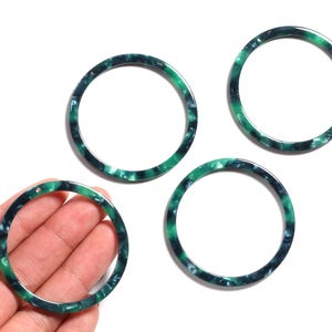 Op de afbeelding: Vier groene en zwarte gemarmerde cirkelhangers van hars op een witte achtergrond. Eén hanger wordt in een hand gehouden. De hangers zijn cirkelvormig met een gat om te rijgen.