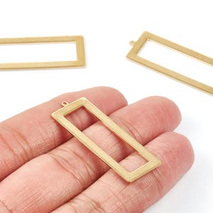 Puede incluir: Tres colgantes rectangulares dorados con un pequeño lazo en la parte superior para sujetar a la joyería. Un colgante se sostiene en una mano. Los colgantes tienen un centro hueco y un acabado liso y mate. El fondo es blanco.