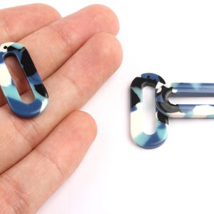 Peut inclure: Deux pendentifs ovales ouverts avec un motif marbré dans les tons de bleu, blanc et noir. Un pendentif est tenu dans une main, tandis que l'autre est positionné à côté. Les pendentifs ont un trou en haut pour l'enfilage.