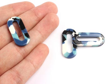 May include: Two open oval-shaped pendants with a marbled pattern in shades of blue, white, and black. One pendant is held in a hand, while the other is positioned next to it. The pendants have a hole at the top for stringing.