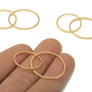 Puede incluir: Componentes de joyería de metal dorado. La imagen muestra varios conjuntos de anillos ovalados entrelazados. Un conjunto se sostiene en una mano. Los anillos son delgados y tienen un acabado liso y pulido. El fondo es blanco.