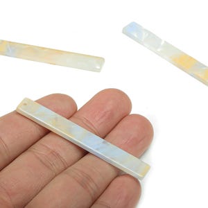 May include: Rectangular, iridescent acrylic beads in shades of white, yellow, and blue. One bead is held in a hand, while two others are scattered nearby. Each bead has a small hole for stringing.