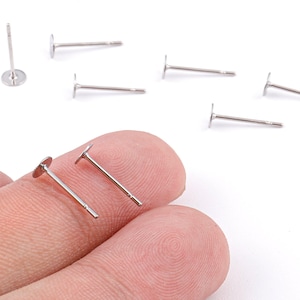 May include: Silver-colored earring posts with flat, round pads. Several posts are scattered on a white surface, with two held between fingers. The posts have a simple, minimalist design, suitable for DIY jewelry making.
