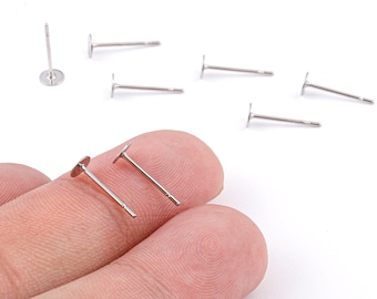 May include: Silver-colored earring posts with flat, round pads. Several posts are scattered on a white surface, with two held between fingers. The posts have a simple, minimalist design, suitable for DIY jewelry making.