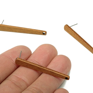 May include: Three wooden bar earrings with a post and a small hole at the end. The earrings are a light brown color and are displayed on a white background. One earring is being held in a person's hand.