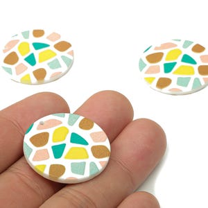 May include: Three round, white polymer clay pendants with a mosaic pattern. The mosaic design features irregular shapes in yellow, teal, brown, and pink. One pendant is held in a hand, and the other two are on a white background.