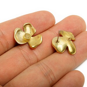 May include: A pair of gold-colored flower-shaped stud earrings. One earring is face up, showing the petals and a textured center. The other earring is face down, showing the post and back. The earrings are on a hand.