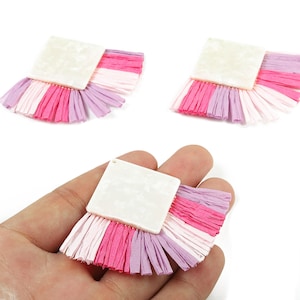 May include: Three square-shaped earrings with a white top and a fringe of pink, white, and purple raffia. One earring is held in a hand. The earrings are set against a white background.