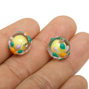 May include: Two clear glass beads with a yellow core and colourful accents. The beads have a clear outer layer with green, orange, and purple details. The beads are held in a hand.