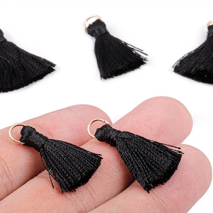 May include: Close-up of several black tassels with gold-colored loops. The tassels are made of a silky material and are displayed against a white background. Two tassels are held in a hand.