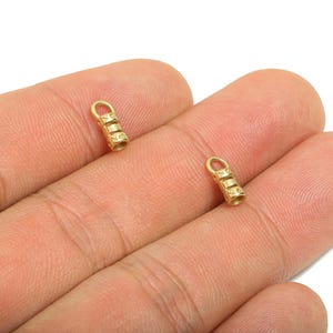 May include: Two small, gold-colored jewelry findings with a cylindrical shape and a loop at the top. The findings have a textured, ribbed design and are shown against a skin-toned background.
