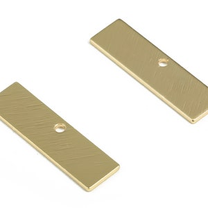 Puede incluir: Dos componentes rectangulares de metal dorado con una textura cepillada. Cada componente tiene un solo agujero centrado. Estos hallazgos se utilizan probablemente en la fabricación de joyas para conectar piezas o como elementos decorativos.