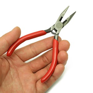 Puede incluir: Primer plano de unos alicates de punta fina con mangos rojos. Los alicates son de metal y tienen un mecanismo de resorte. La herramienta se sostiene en una mano sobre un fondo blanco. Los alicates se utilizan para agarrar, doblar y cortar.