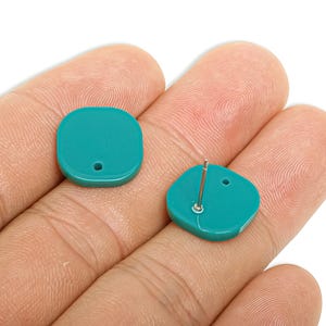 May include: Two teal-colored, square-shaped stud earrings. One earring has a post for insertion, while the other is a matching stud. The earrings are held in a hand against a white background. The earrings are a solid color.