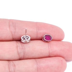 Può includere: Un paio di orecchini a lobo con pietre preziose rosso scuro di forma ovale incastonate in metallo color argento. Gli orecchini sono mostrati su uno sfondo chiaro, evidenziando il loro design e colore. Il perno di un orecchino è visibile.