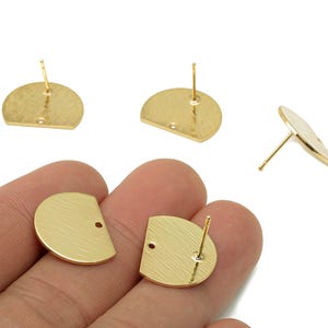 Pode incluir: Brincos de botão dourados em forma de semicírculo com superfície texturizada. Os brincos têm um pino para inserção e um pequeno orifício para fixação. Vários brincos são exibidos, alguns sendo segurados na mão.