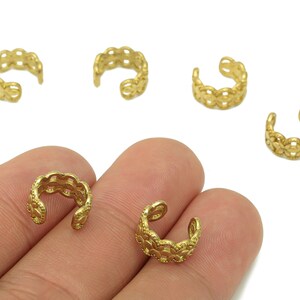 May include: Gold-colored ear cuffs with a delicate, openwork design. The ear cuffs are shown in a group, with some resting on a hand. The openwork pattern creates a scalloped edge.