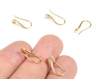 May include: Several gold-colored ear hooks are displayed against a white background. The ear hooks have a curved, open design. One pair of ear hooks is held by a person's fingers. The hooks are made of a shiny, metallic material.