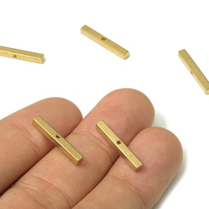 Peut inclure: Gros plan de plusieurs petites barres métalliques rectangulaires dorées avec un seul trou au centre de chacune. Les barres sont posées sur une surface de couleur claire. Ce sont probablement des composants de fabrication de bijoux.